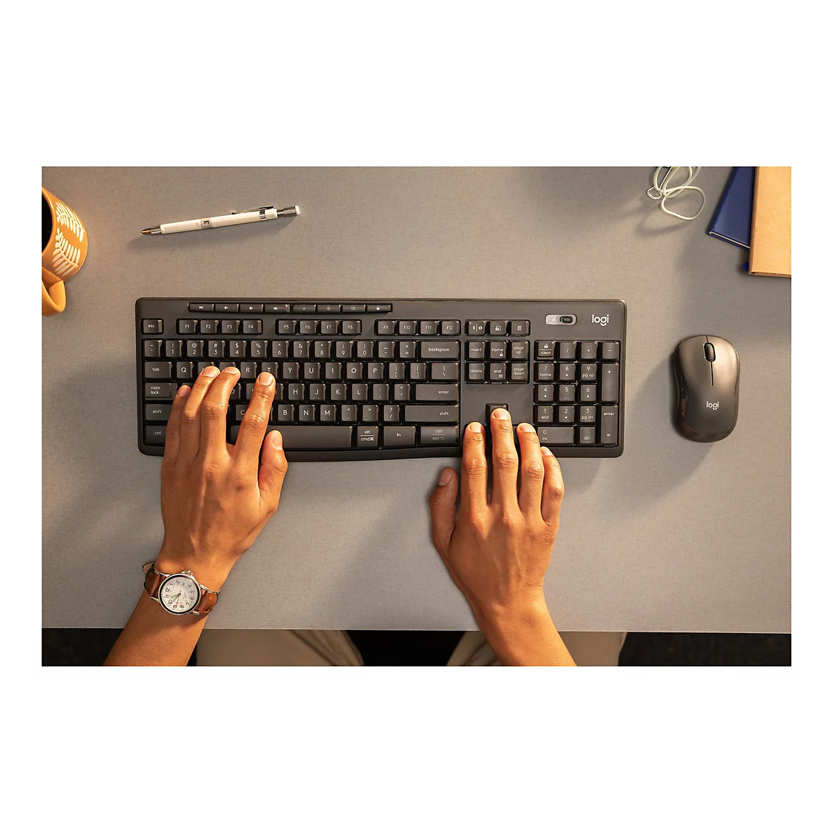 Draufsicht auf eine schwarze Logitech-Tastatur und -Maus, die auf einem grauen Schreibtisch liegen. Die Hände einer Person ruhen auf der Tastatur, eine Uhr am linken Handgelenk.