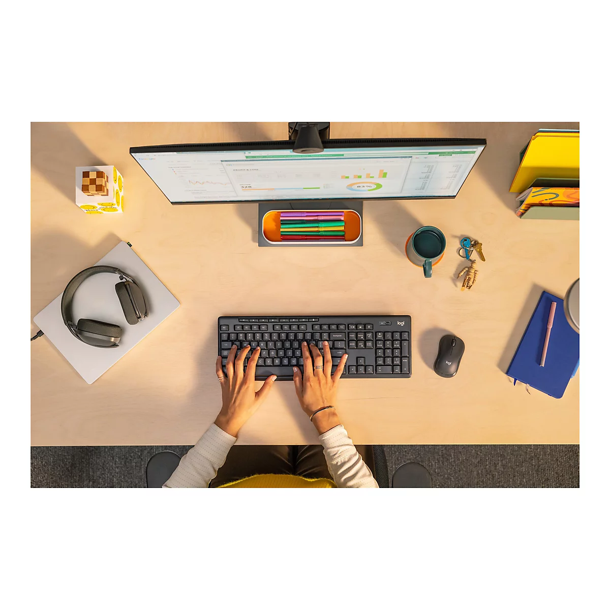 Schreibtischansicht von oben. Eine Person tippt auf einer schwarzen Logitech-Tastatur, daneben eine schwarze Maus. Auf dem Schreibtisch: Monitor, Kopfhörer, Notizbuch, Tasse.