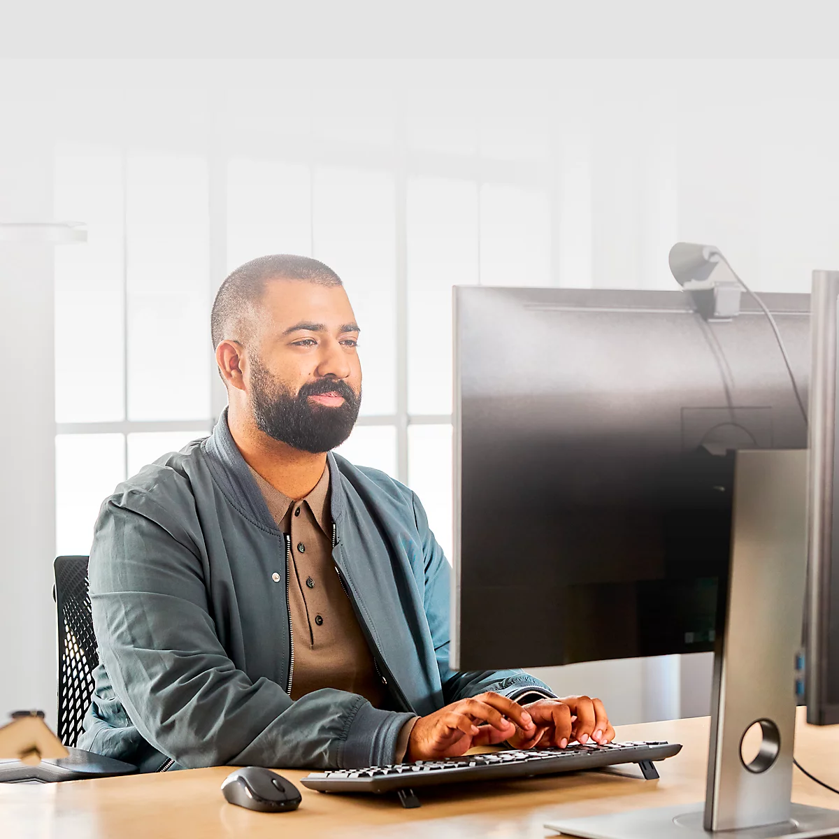 A man with a beard sits at a desk in front of a computer typing. He is smiling slightly.