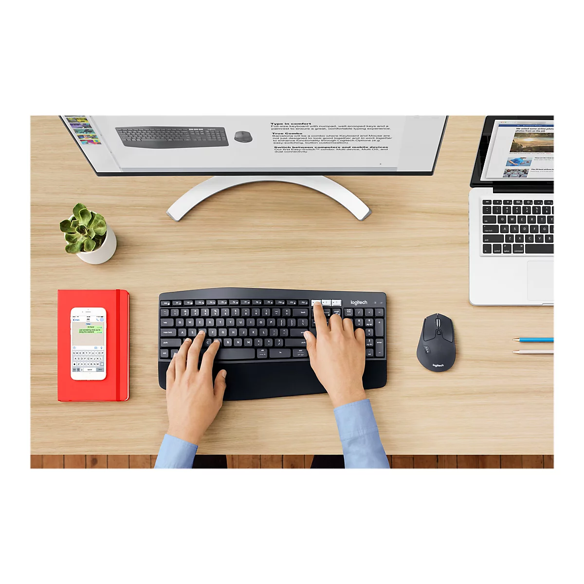 Configuration de bureau avec clavier, souris et smartphone noirs. Des informations s'affichent à l'écran. À côté, un carnet rouge, un moniteur et une petite plante en pot.