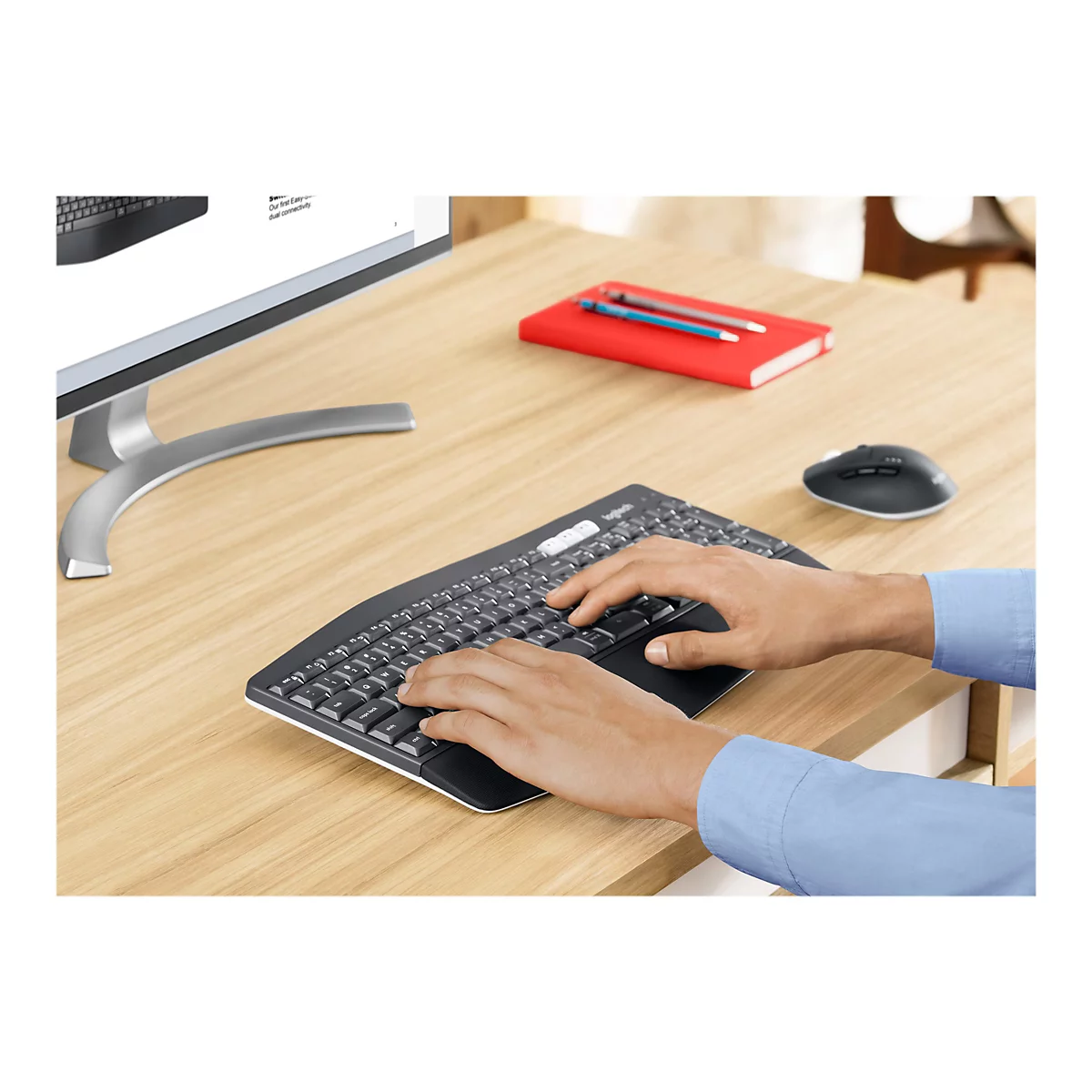 Une personne tape sur un clavier ergonomique. Un cahier rouge, une souris et un moniteur sont sur le bureau.