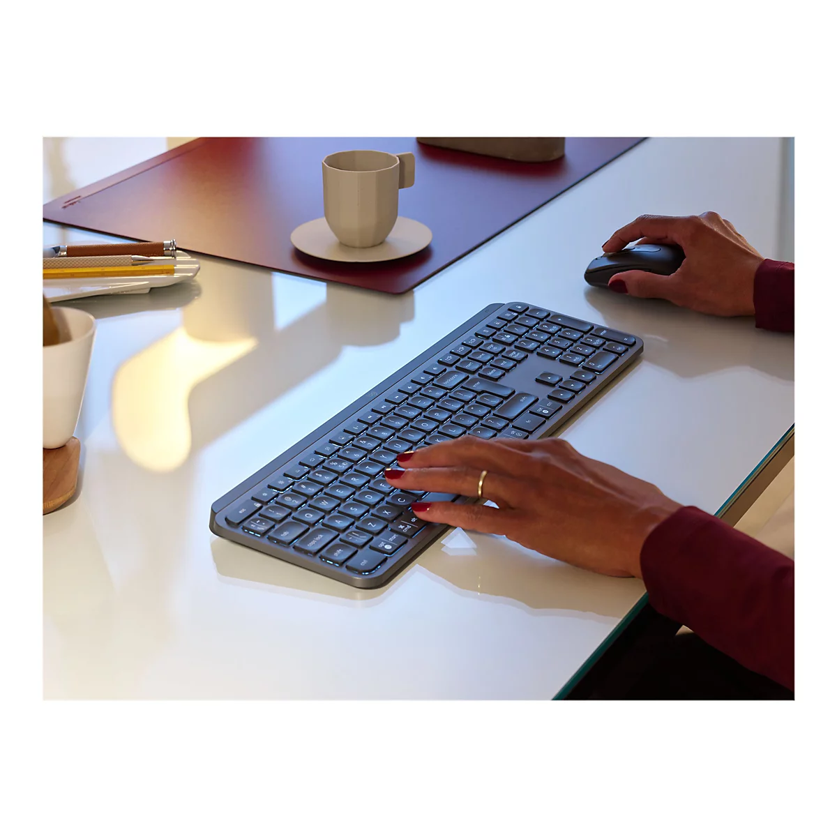 Person, die eine graue Tastatur auf einem weißen Tisch benutzt; daneben liegt eine Maus. Auf dem Tisch steht eine Tasse Kaffee.