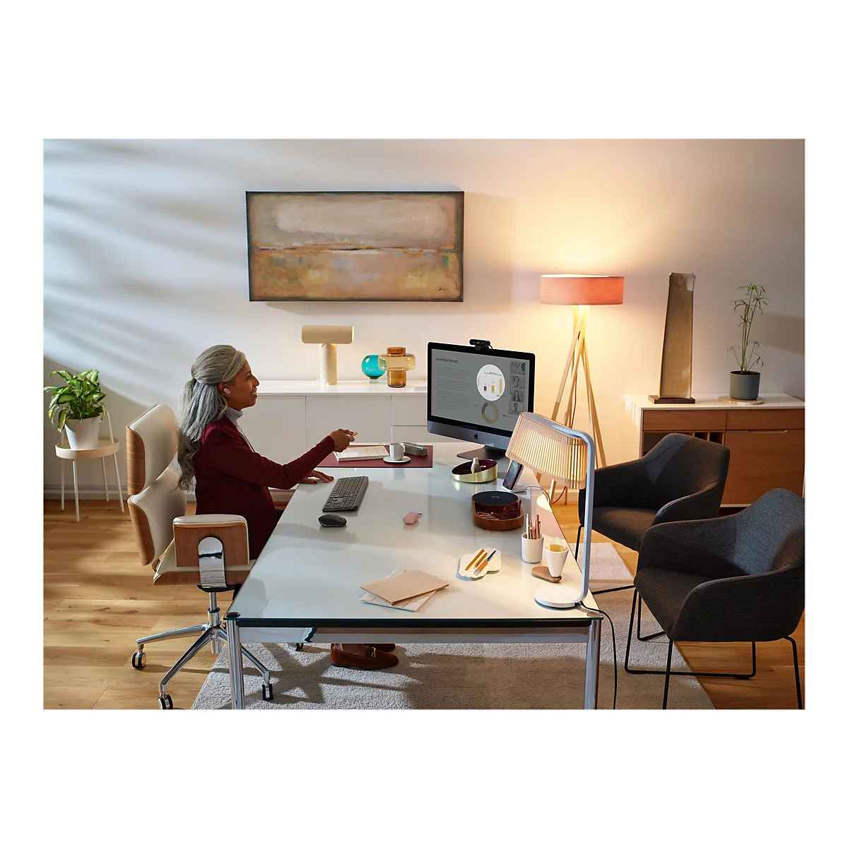 Eine Frau sitzt an einem Schreibtisch und telefoniert. Auf dem Monitor sind Diagramme zu sehen. Es gibt eine Schreibtischlampe, eine Stehlampe und ein Kunstwerk an der Wand.