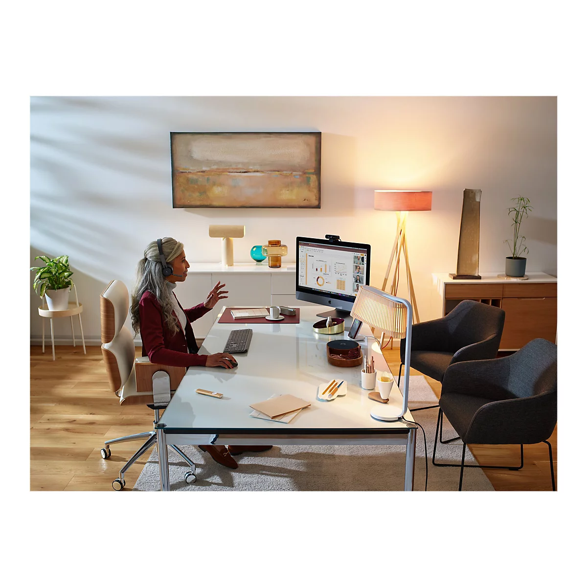Eine Frau mit Headset sitzt an einem Schreibtisch und nimmt an einer Videokonferenz teil. Auf dem Schreibtisch befinden sich ein Monitor, Papierkram und eine kleine Lampe.