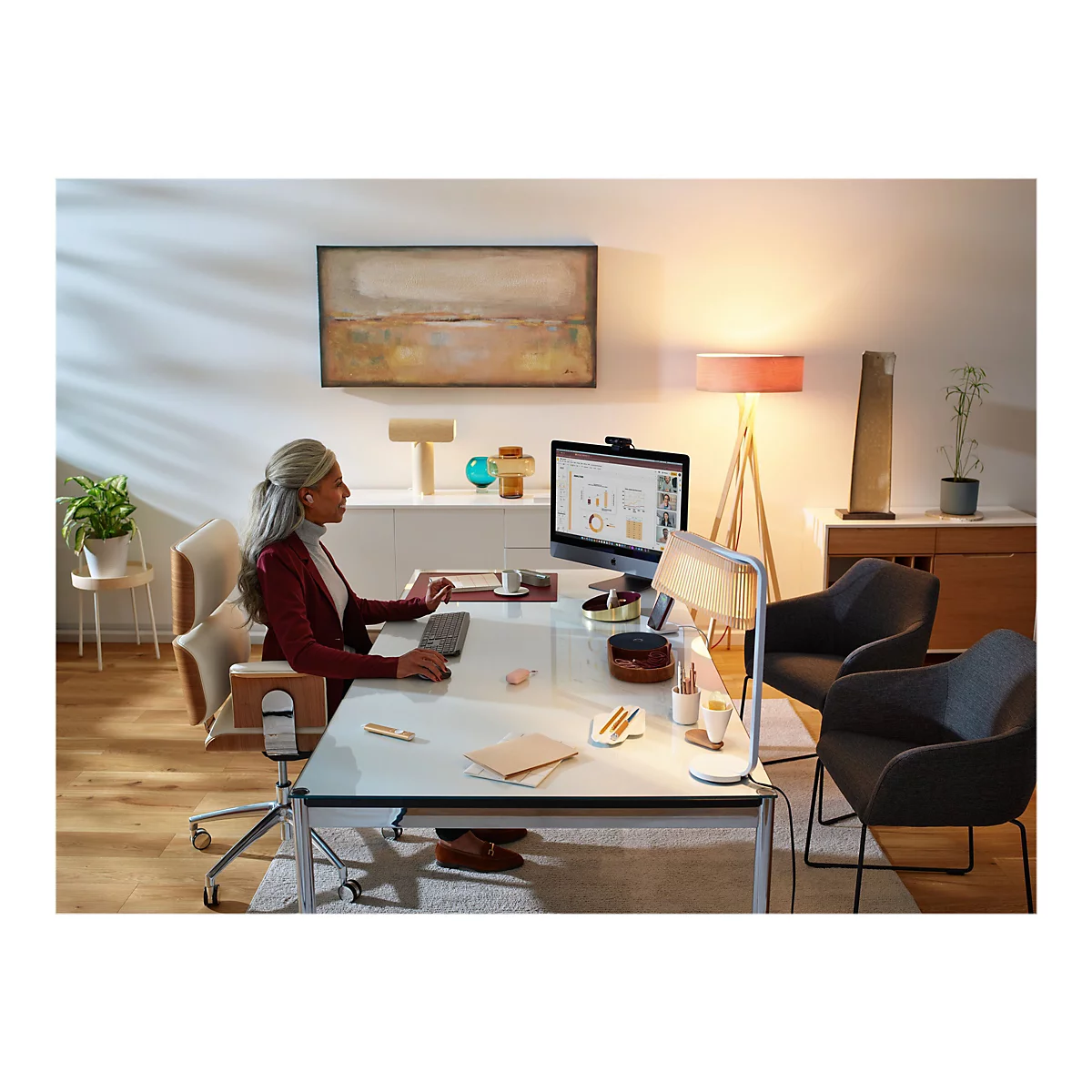 Eine Frau mit grauen Haaren sitzt an einem Schreibtisch. Sie trägt ein rotes Jackett und Kopfhörer. Auf dem Bildschirm des Computers ist ein Diagramm zu sehen.