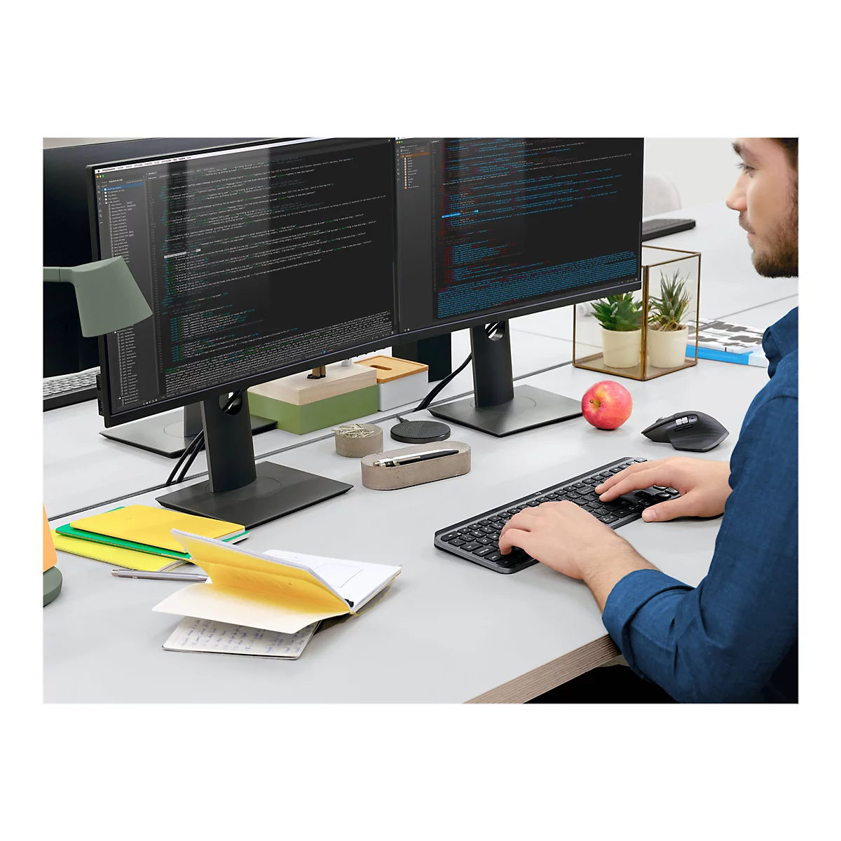 Ein Mann tippt auf einer Tastatur vor zwei Monitoren, die Code zeigen. Auf dem Schreibtisch liegen eine Maus, ein Apfel, Notizen und Dekorationsartikel.