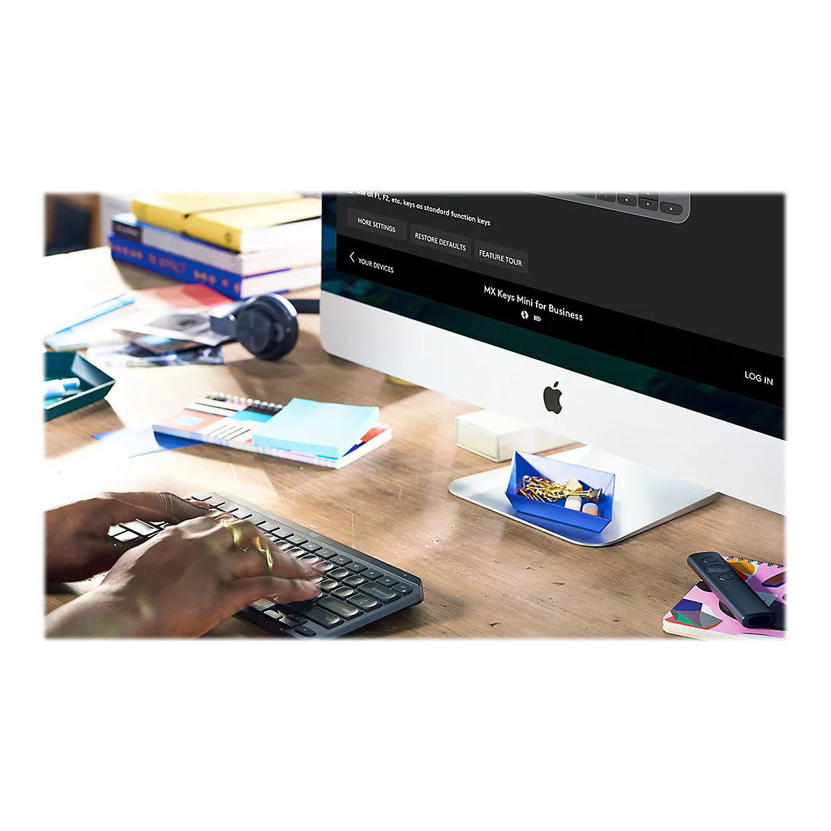 Person tippt auf Tastatur vor Apple-Monitor. Auf dem Tisch liegen KopfhÃ¶rer, Notizen, und Accessoires.