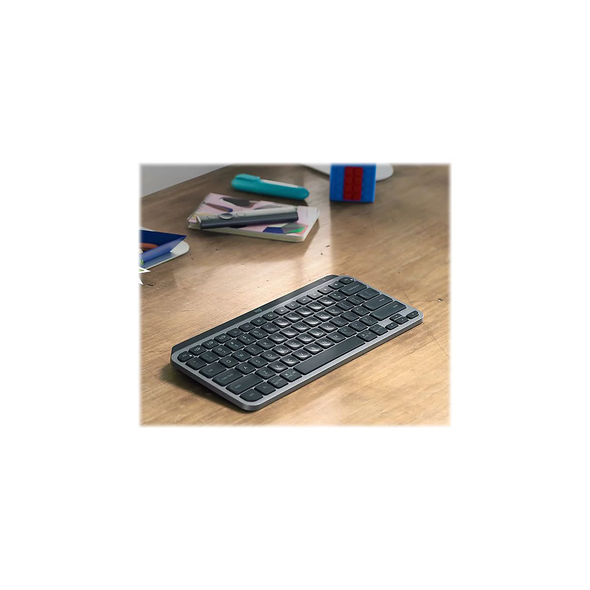 Drahtlose Tastatur auf einem Holztisch mit Notizbuch, Stift und Spielzeug im Hintergrund.