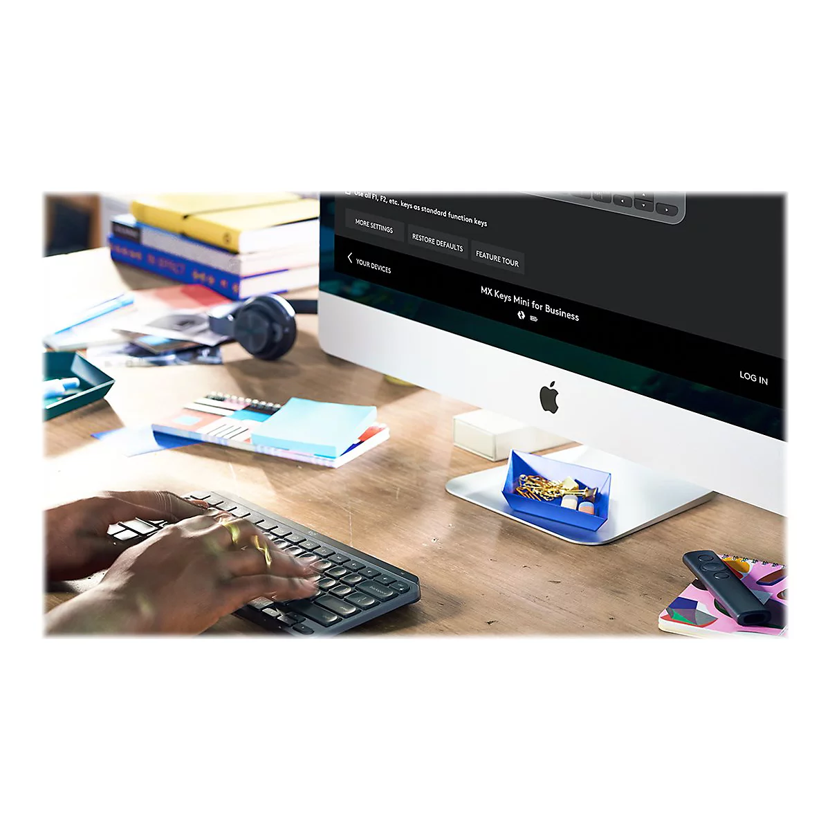 Person tippt auf einer Tastatur, vor einem Apple-Monitor, in dem ein geöffnetes Menü angezeigt wird. Auf dem Schreibtisch liegen Büroartikel.