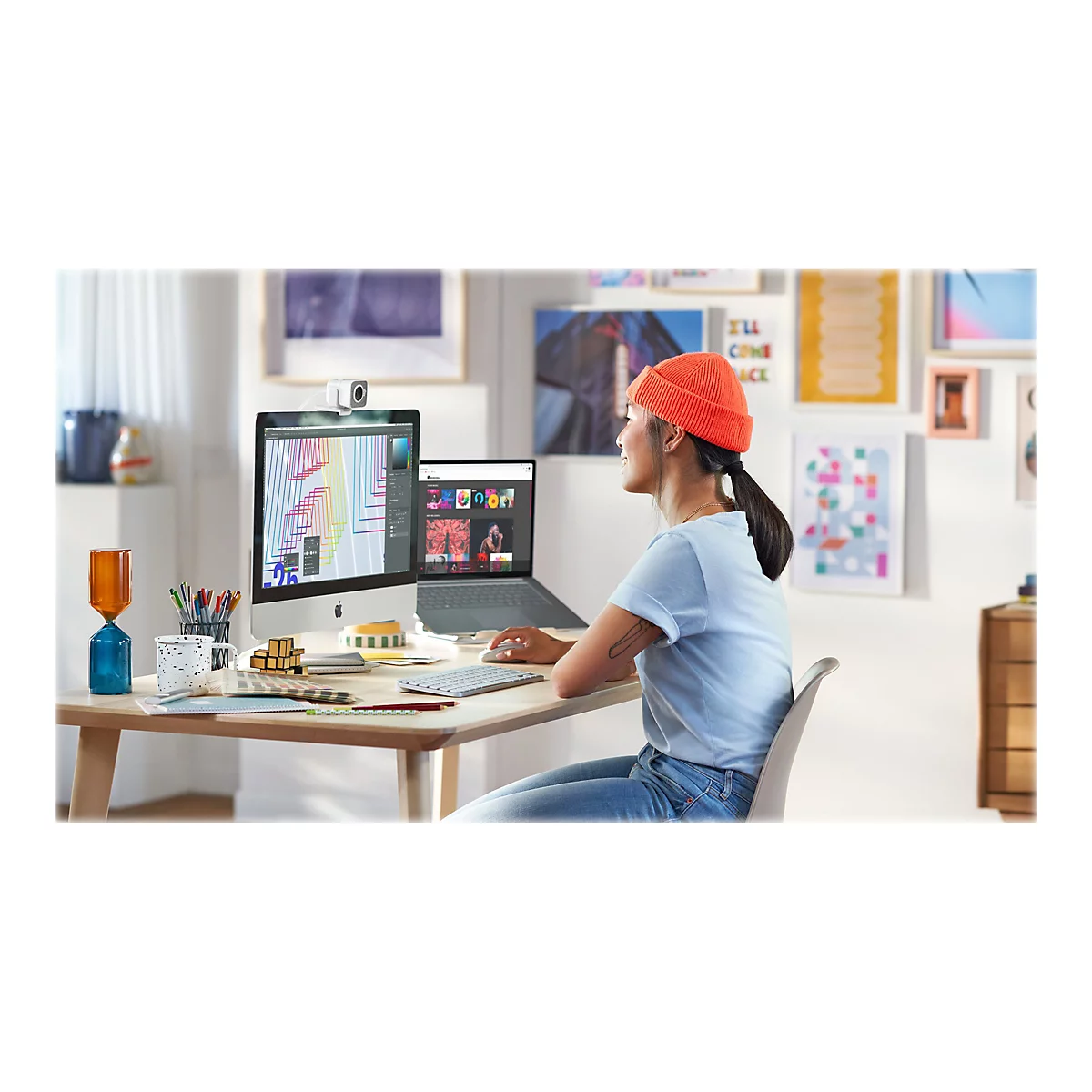 Junge Frau sitzt an einem Schreibtisch mit Computer und Laptop. Sie trägt eine orangefarbene Mütze und schaut lächelnd zur Seite.