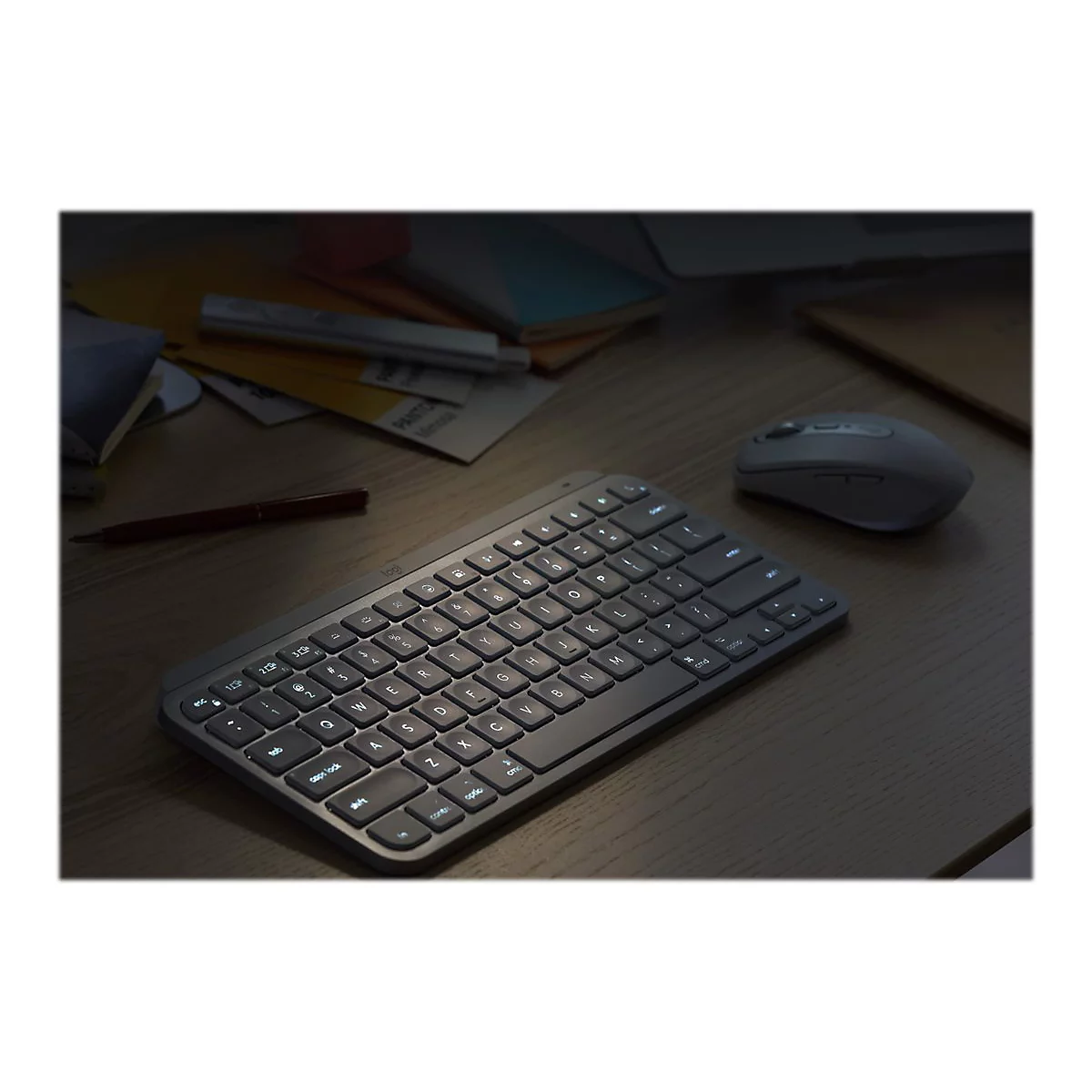 Draufsicht auf eine beleuchtete Tastatur und Maus auf einem Schreibtisch. Im Hintergrund liegen Notizen und ein Stift.