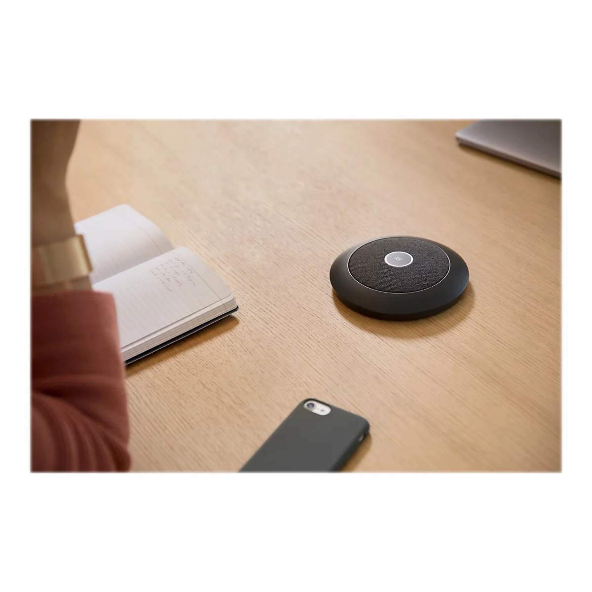 Rundes schwarzes Konferenzmikrofon auf einem Holztisch neben einem Notizbuch und Smartphone.