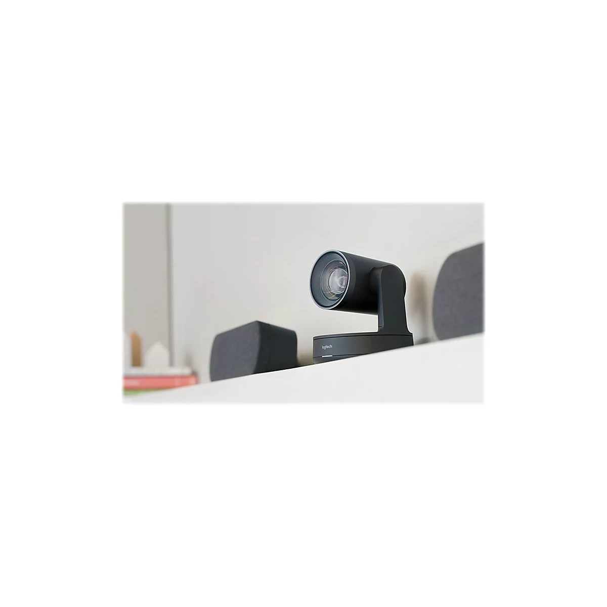 Schwarze Logitech-Webcam auf einer weißen Ablage. Im Hintergrund: Graue Lautsprecherboxen und ein Buch.