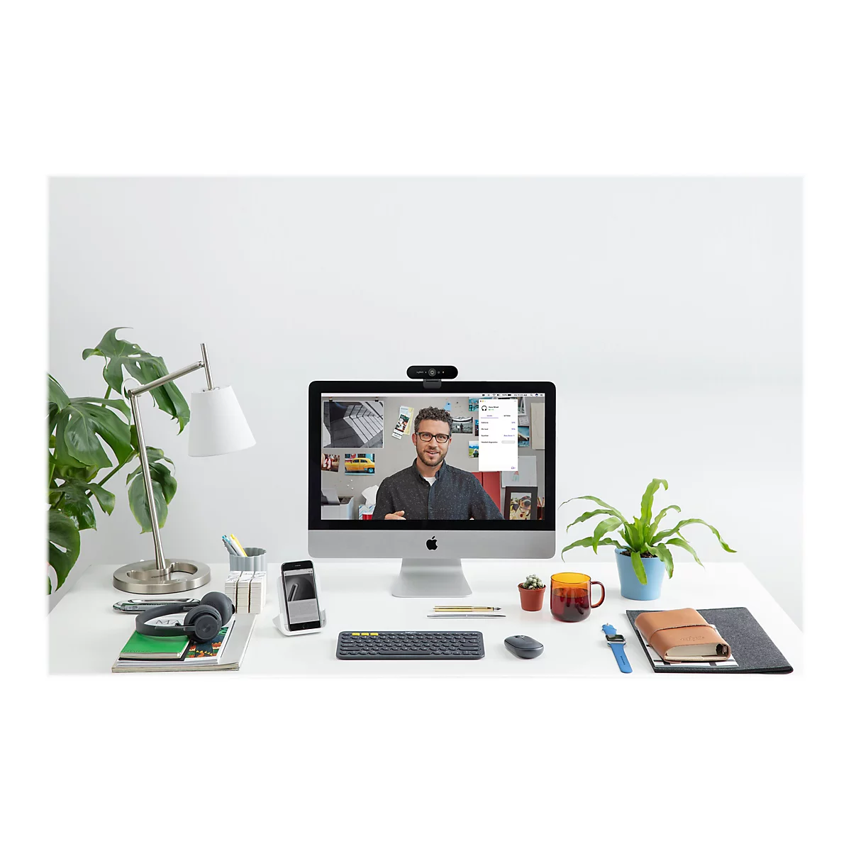 Un espace de travail avec un Ã©cran affichant une visioconfÃ©rence. Des plantes, des cahiers, des stylos, un casque et une webcam sont sur la table.