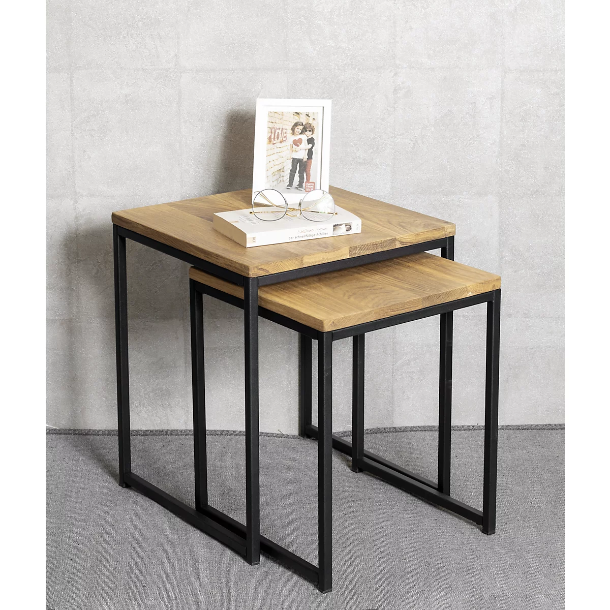 Deux tables carrées en bois avec des cadres métalliques sont empilées l'une sur l'autre. Sur la table du dessus se trouvent un livre, des lunettes et un cadre photo.