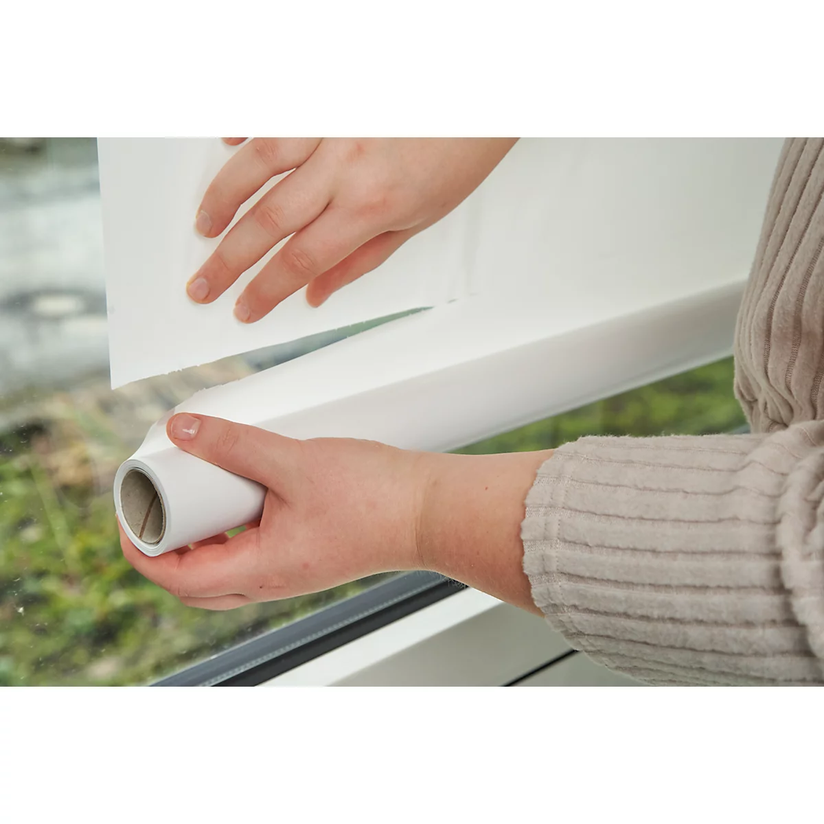 Hände halten eine Rolle Fensterfolie und ein Stück davon in der Hand. Beige Pullover, Fensterrahmen.