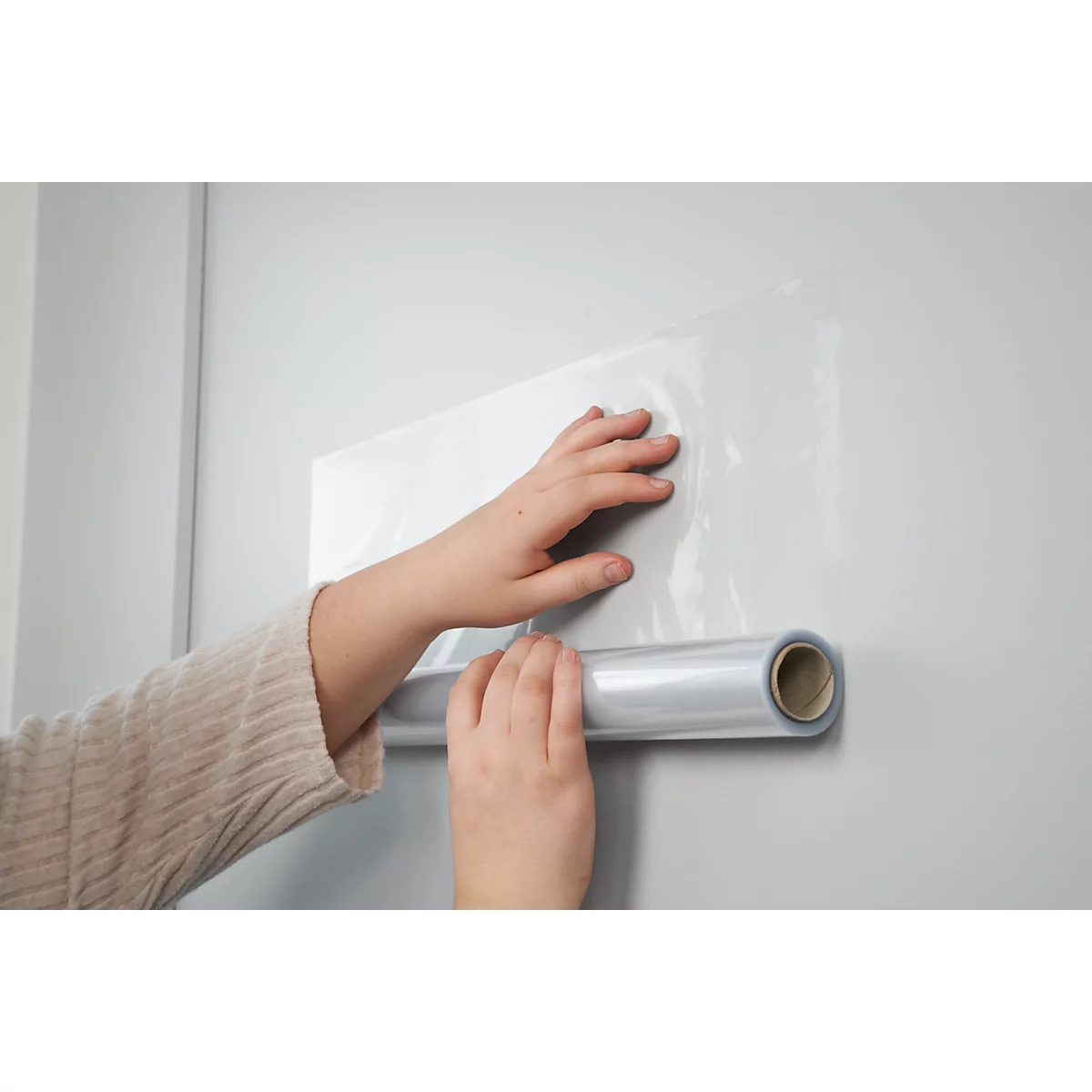 Dos manos sosteniendo una lámina transparente, desenrollándose de un rollo, sobre una pared blanca.