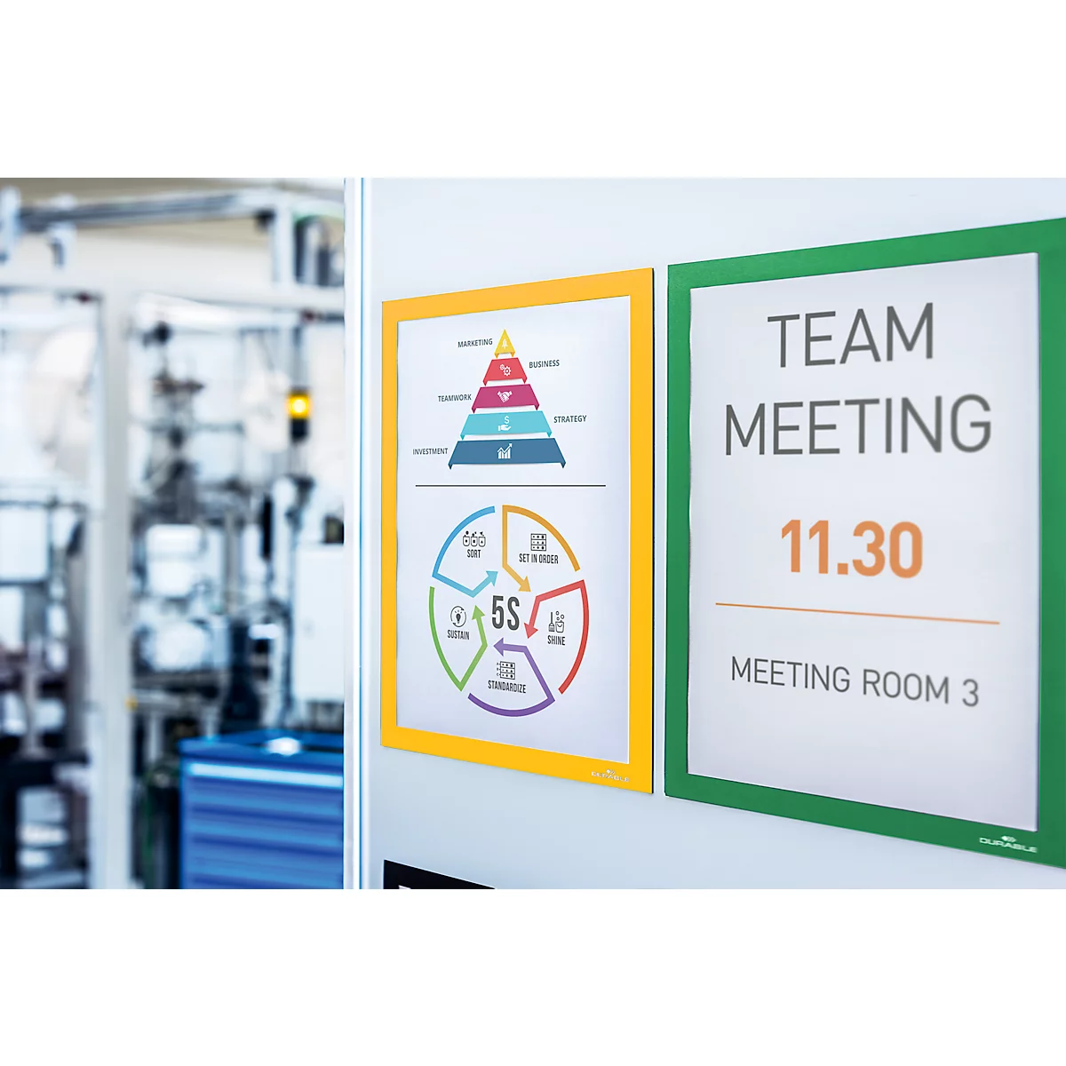 Two signs are mounted on a white background. On the left, a yellow-framed diagram. On the right, a green sign with the words TEAM MEETING, 11.30, Meeting Room 3.