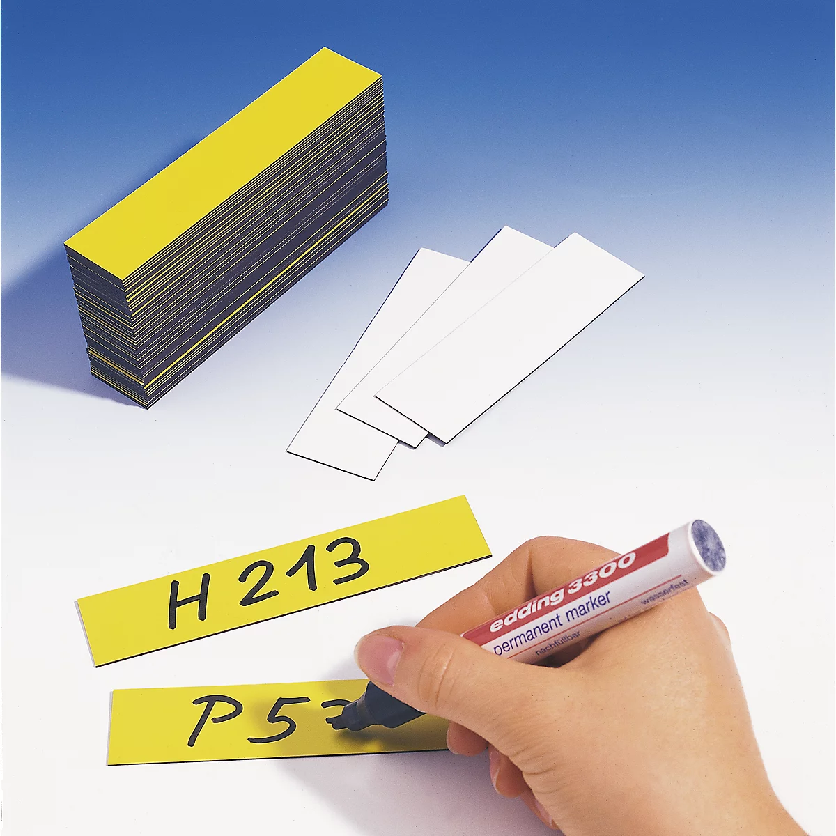 A hand writes on yellow labels with an Edding marker. Numbers and letters can be seen on the labels. A stack of yellow labels is in the background.