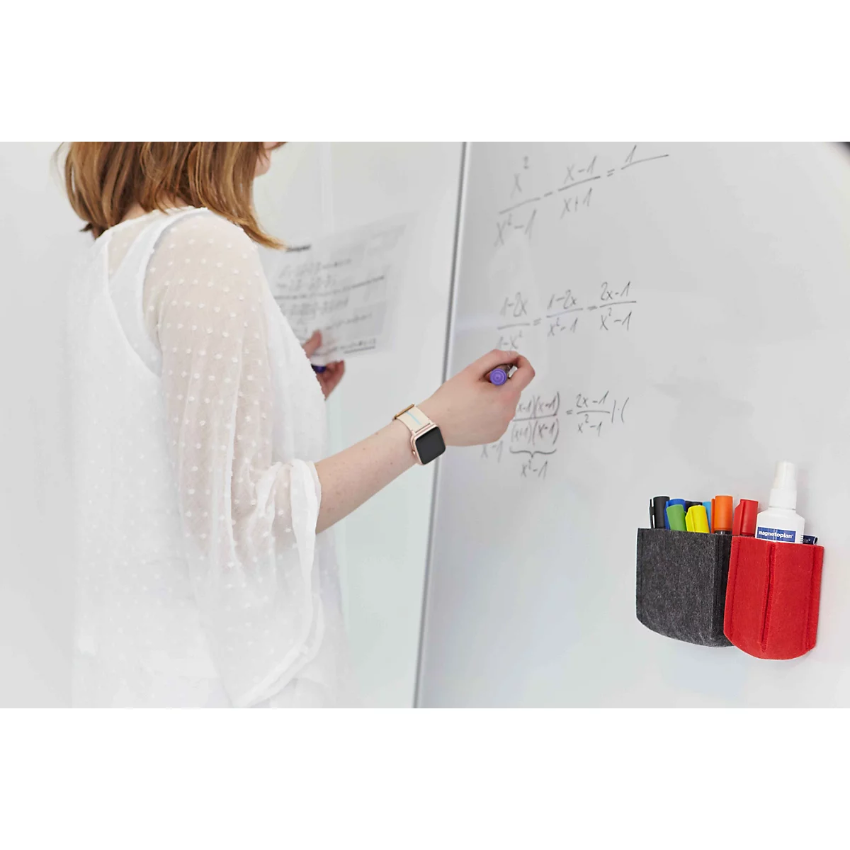 Une femme écrit des formules mathématiques sur un tableau blanc avec un stylo, des pochettes en feutre pour stylos sont accrochées à côté.