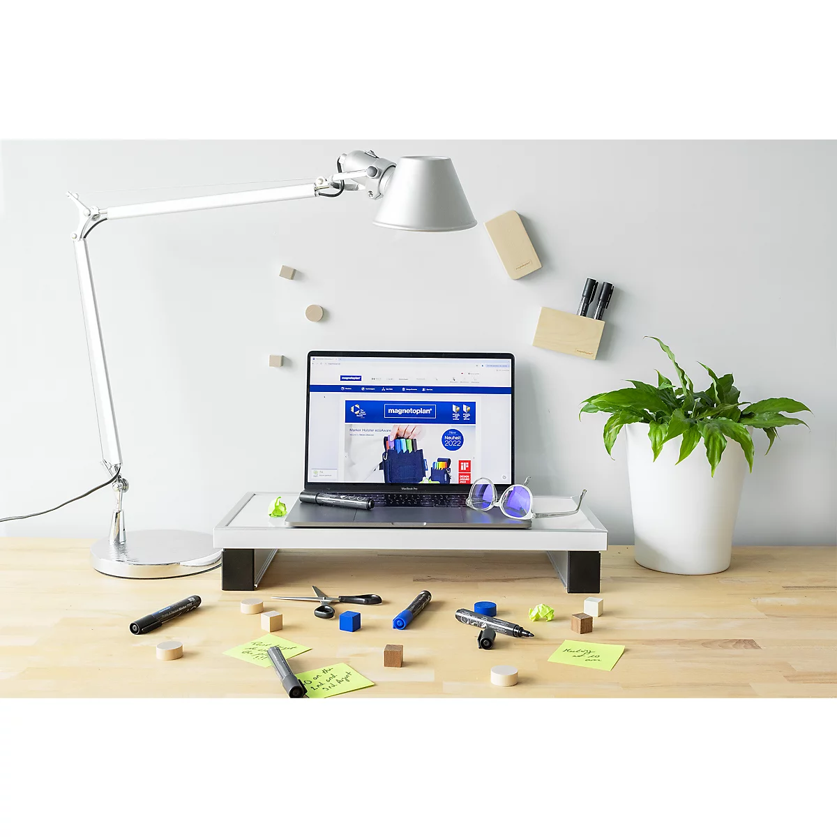 Laptop auf einem Monitorständer auf einem Schreibtisch mit Schreibtischlampe, Pflanze und verstreutem Büromaterial.