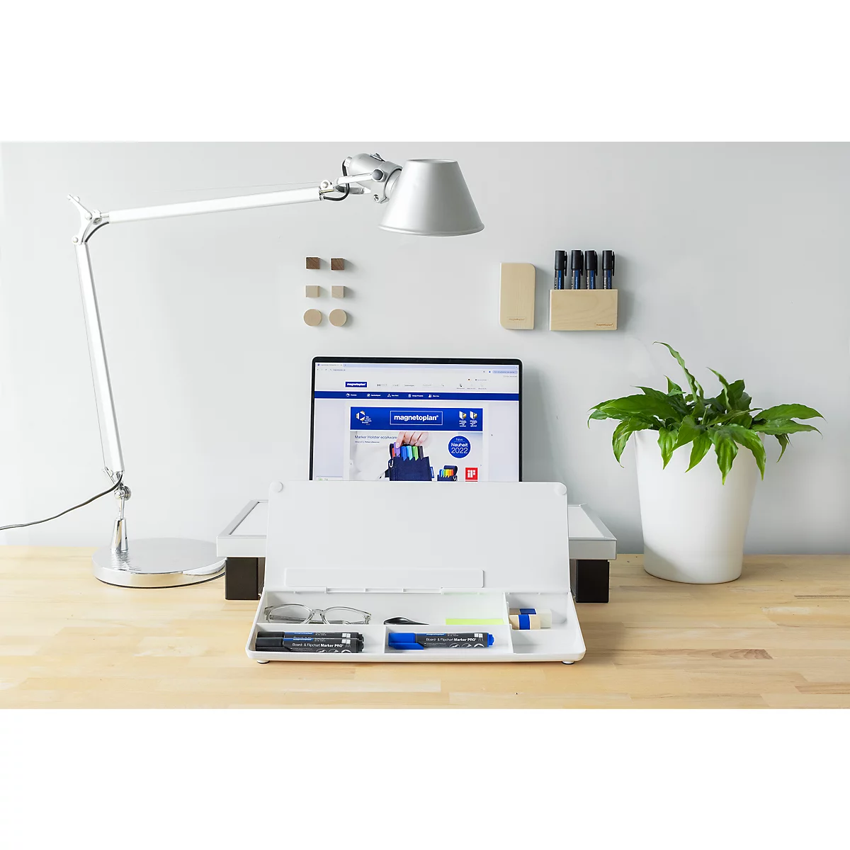 Aufgeräumter Schreibtisch mit weißem Organizer, Lampe, Laptop und Pflanze vor einer hellen Wand.