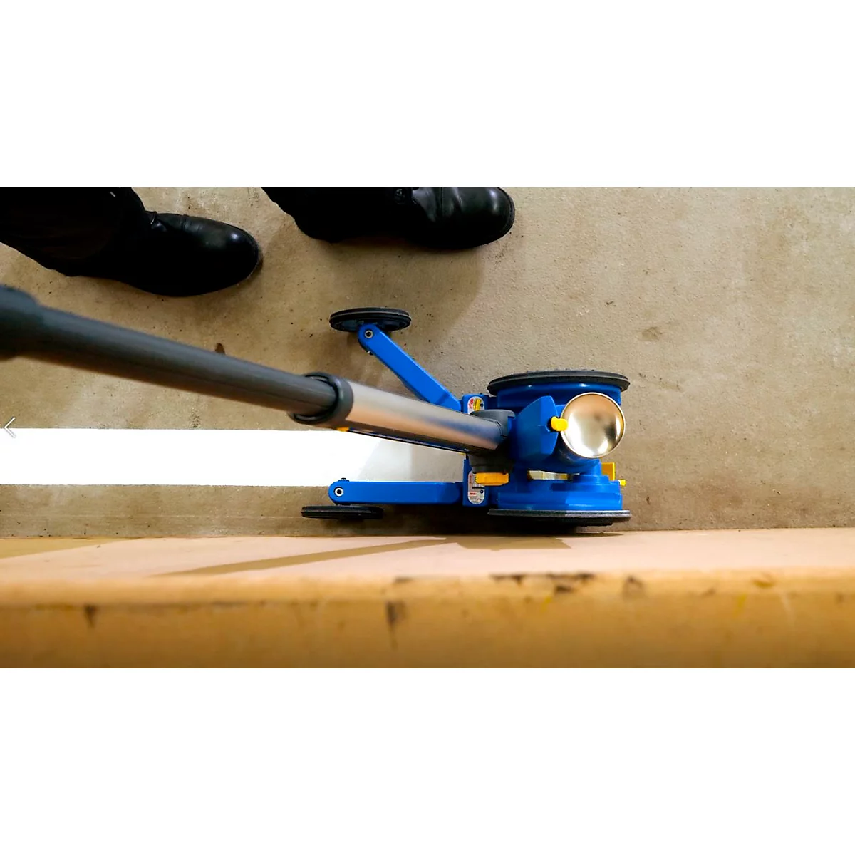 Dispositivo azul levantando un panel de madera. Persona con zapatos negros cerca. Luz brillante.
