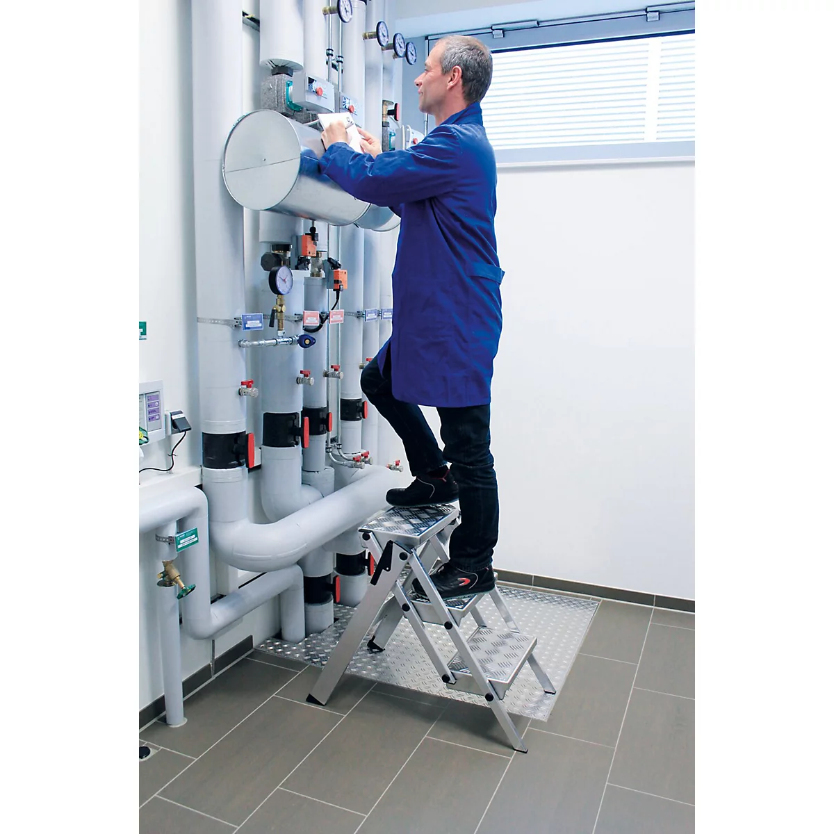 Un homme en veste bleue se tient sur un escabeau pour travailler sur des tuyaux. Tuyaux et instruments de mesure en arrière-plan.