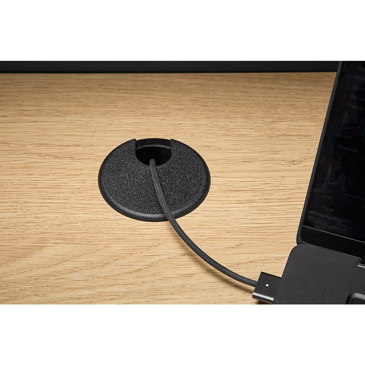 Close-up van een zwarte kabeldoorvoer op een houten oppervlak. Een kabel loopt door de opening. Daarnaast een laptop.