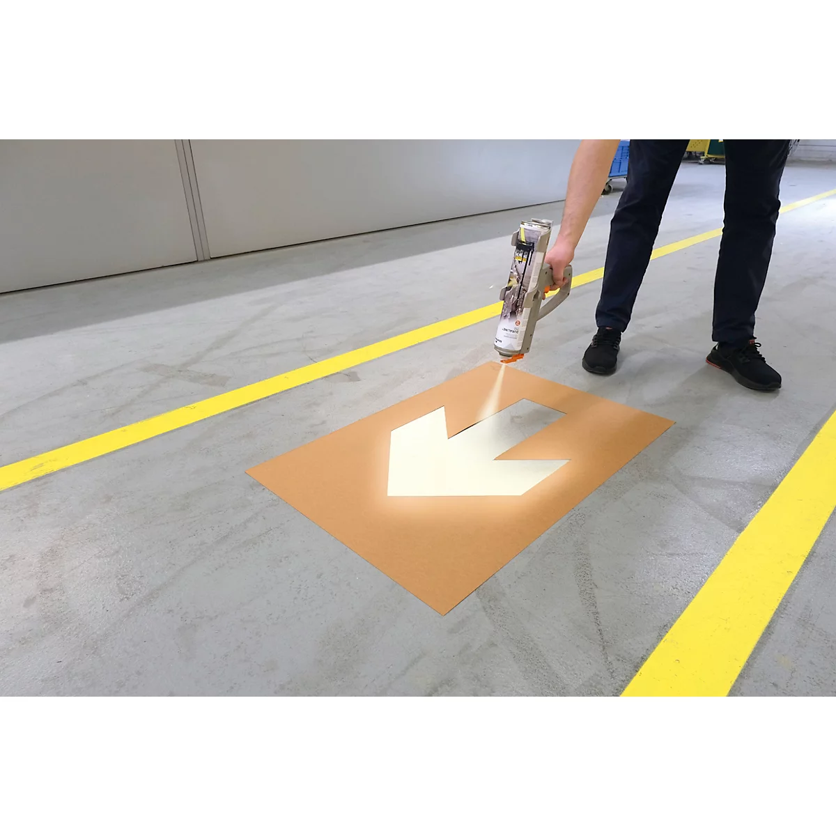 Person besprüht mit einer Sprühpistole einen Pfeil auf dem Boden. Gelbe Markierungen am Boden.