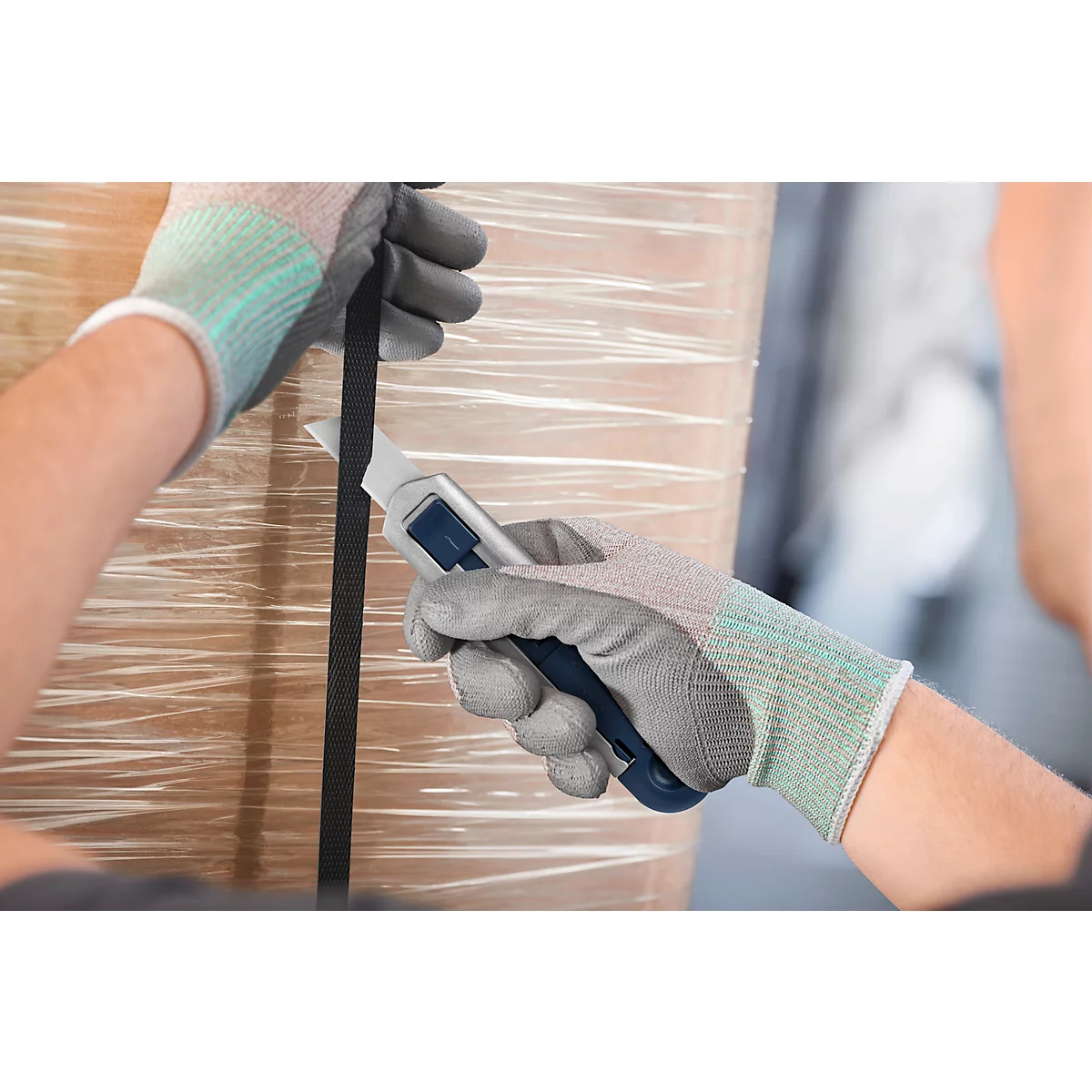 Person mit Arbeitshandschuhen schneidet Verpackung mit einem Messer auf. Die Hände halten Messer und Band.