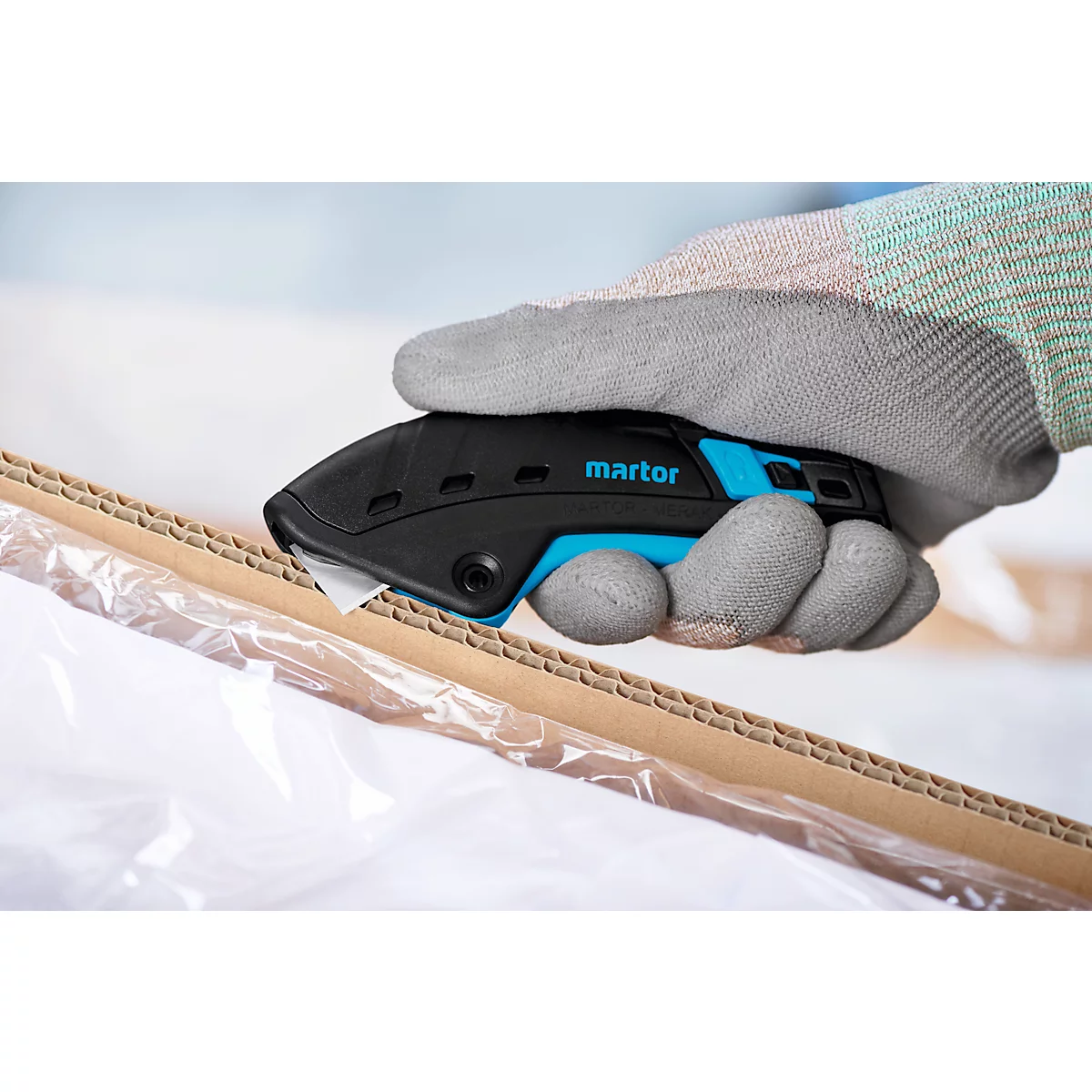 Hand mit grauem Handschuh hält ein schwarzes Cutter-Messer mit blauen Akzenten, schneidet Pappe.
