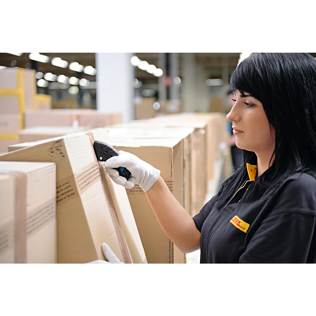 Eine Frau in DHL-Uniform scannt Pakete in einem Lager, sie trägt Handschuhe und blickt konzentriert.