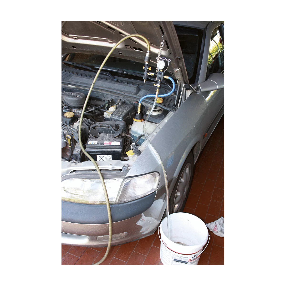 Motor eines silbernen Autos mit geöffneter Motorhaube, verbunden mit Schläuchen und einem Messgerät, Eimer daneben.