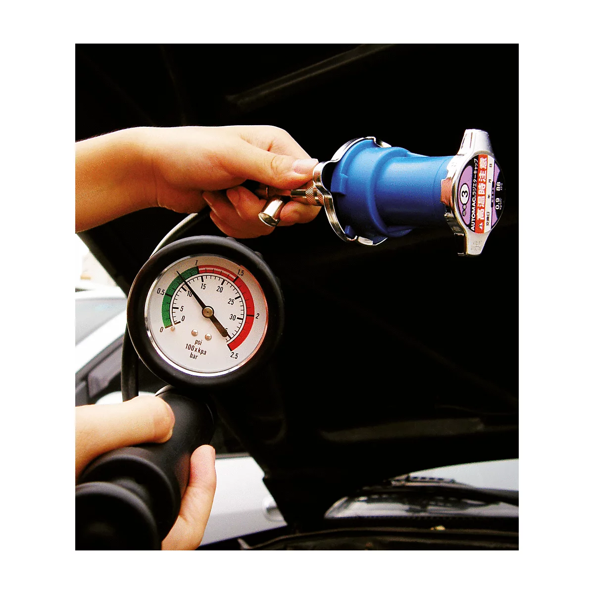 Person hält ein Kühlsystem-Prüfgerät über dem Motorraum eines Autos, ein Manometer zeigt den Druck an.