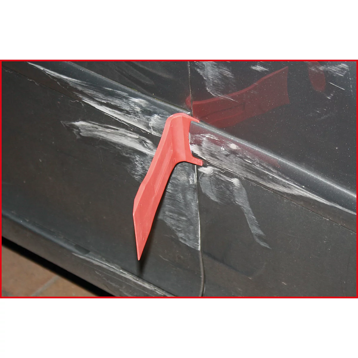 Ein rotes Werkzeug an einer beschädigten Autotür mit Kratzern und weißen Markierungen. Detailaufnahme.