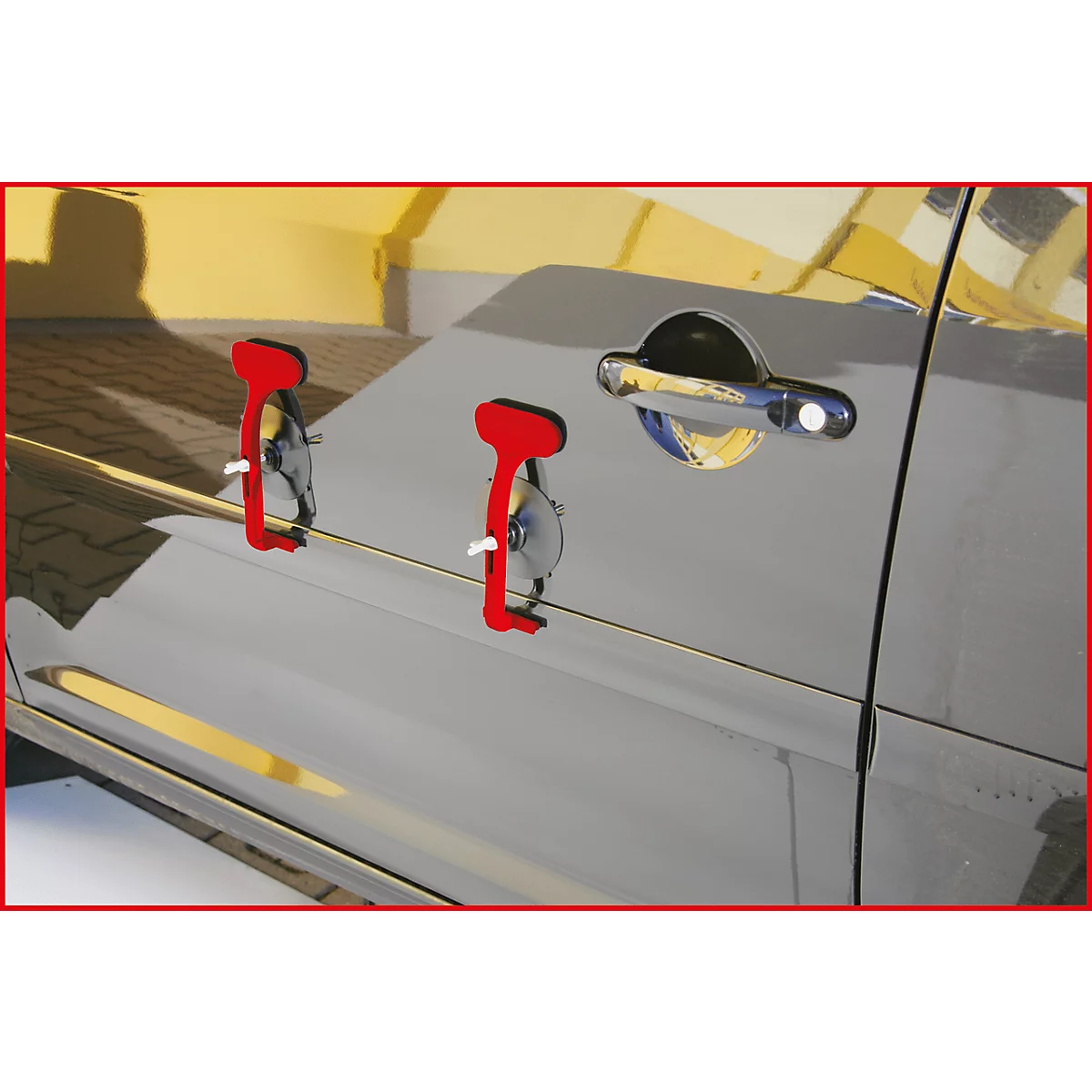 Rote Ausbeulwerkzeuge an Autotür. Im Hintergrund die gelbe Fassade eines Gebäudes.