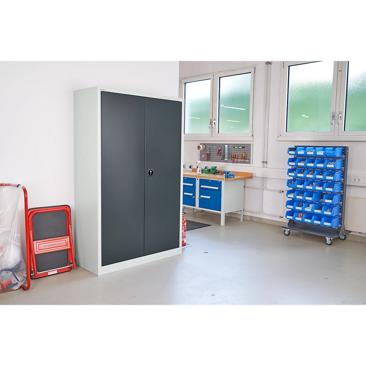 Grijze metalen kast met dubbele deuren in werkplaats naast rode opvouwbare opstap. Werkbank en schappen op de achtergrond.