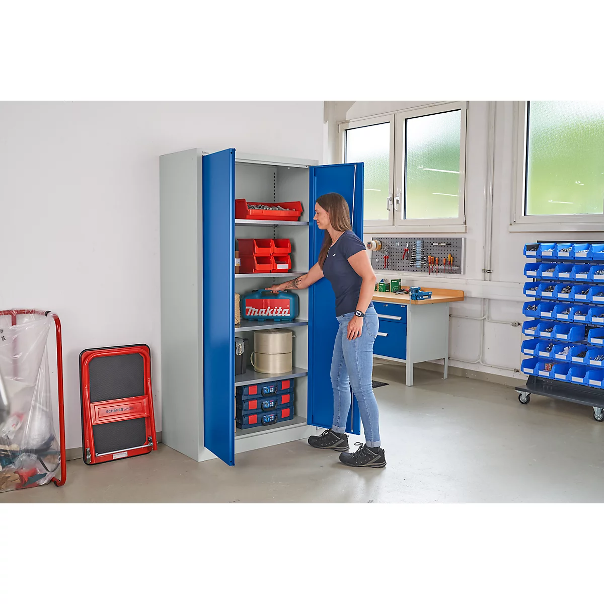 Een vrouw opent een blauwe kast in een werkplaats. Binnenin staan gereedschapskisten en accessoires.