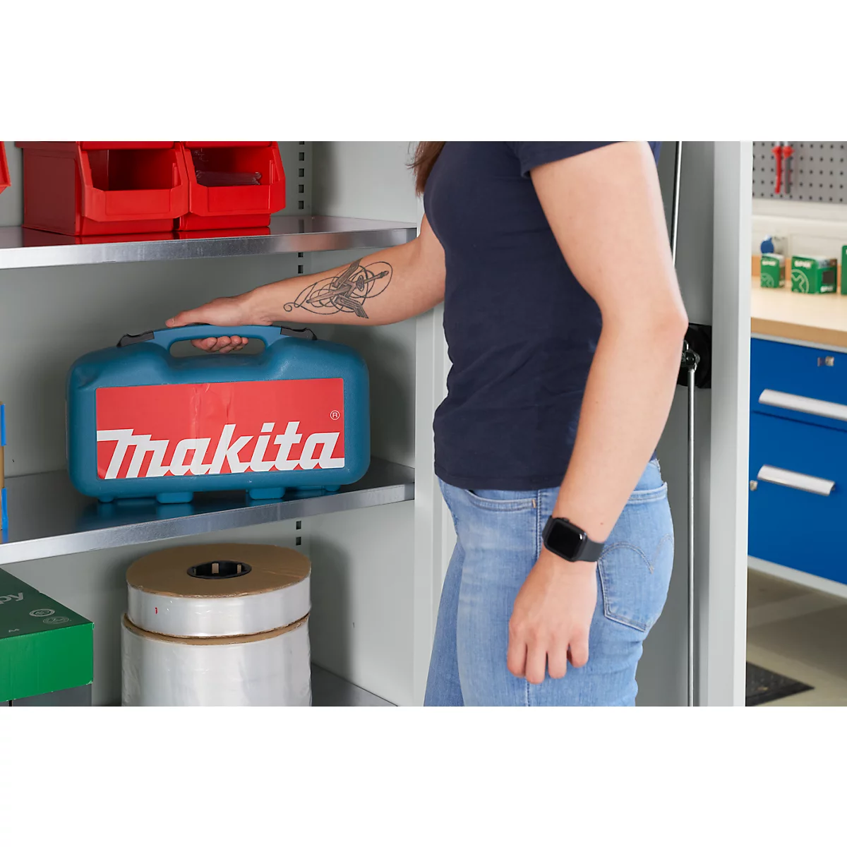 Een persoon in een spijkerbroek en een donkerblauwe t-shirt staat bij een kast. Op de plank ligt een blauwe Makita gereedschapskist.