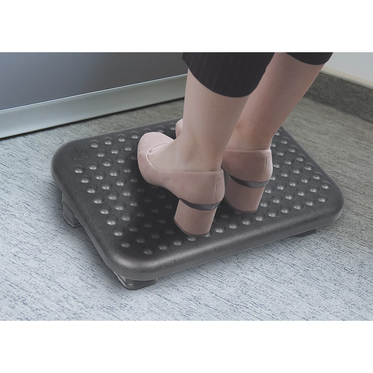 Füße in beigen Schuhen stehen auf einer schwarzen, genoppten Fußstütze unter einem Schreibtisch.