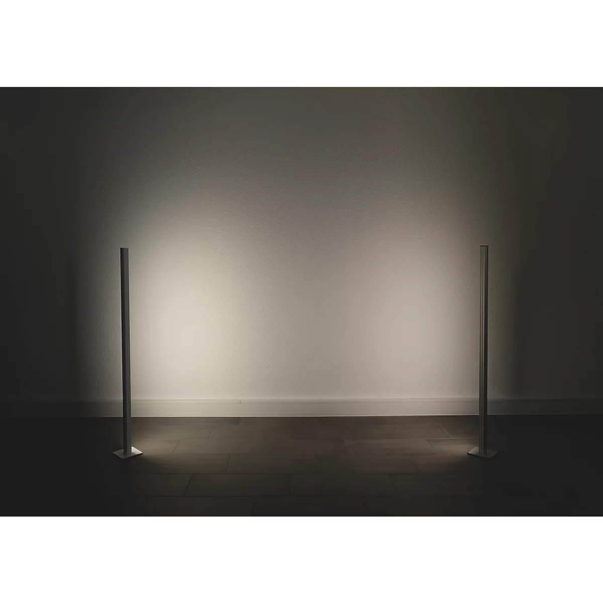 Zwei schlanke, silberfarbene Stehleuchten stehen vor einer hellen Wand. Die Lampen beleuchten den Boden.
