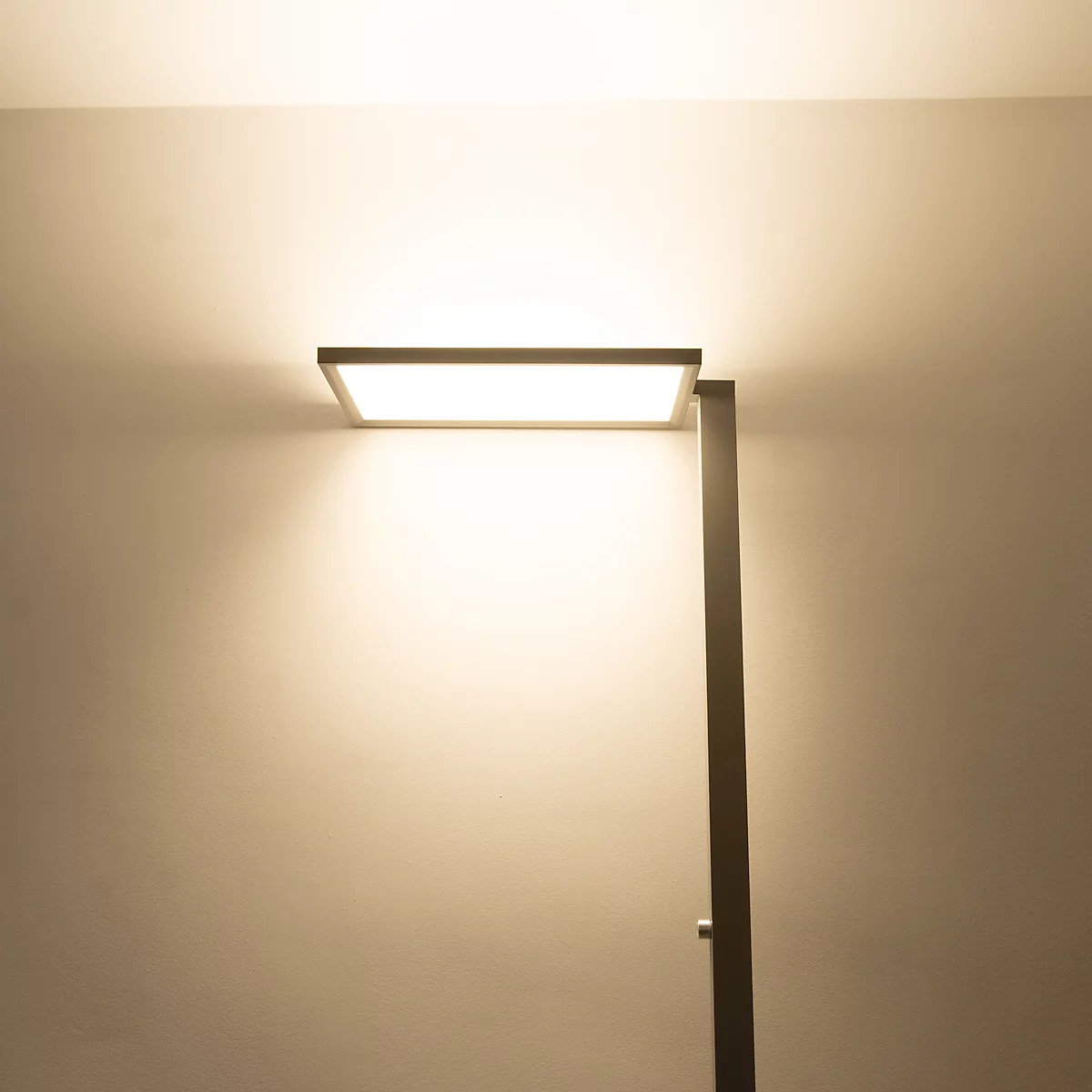 Moderne Stehleuchte mit flachem, rechteckigem Leuchtkopf, der warmes Licht an die Wand strahlt.