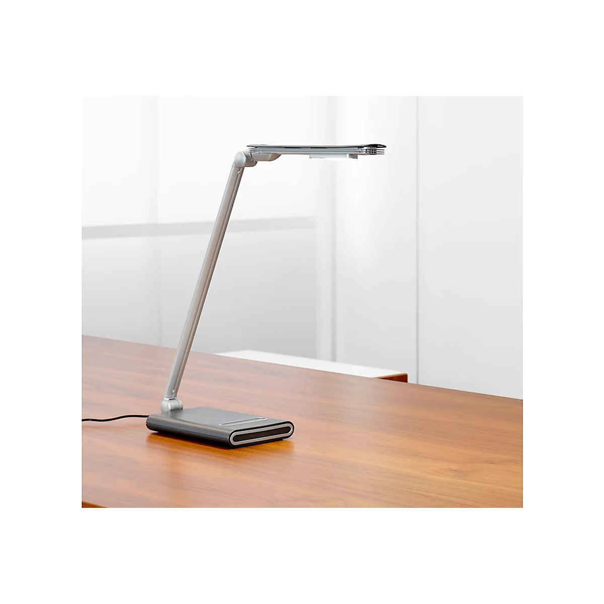 Modern, silver desk lamp on a brown wooden surface. The lamp head is rectangular. In the background a white room.