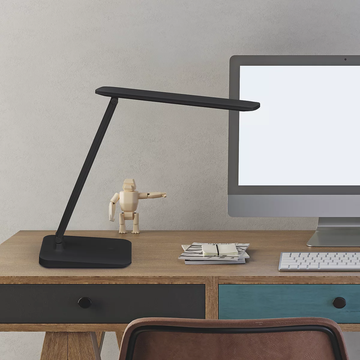 Schwarze Schreibtischlampe auf Holzschreibtisch. Daneben Computer und Holzpuppe.