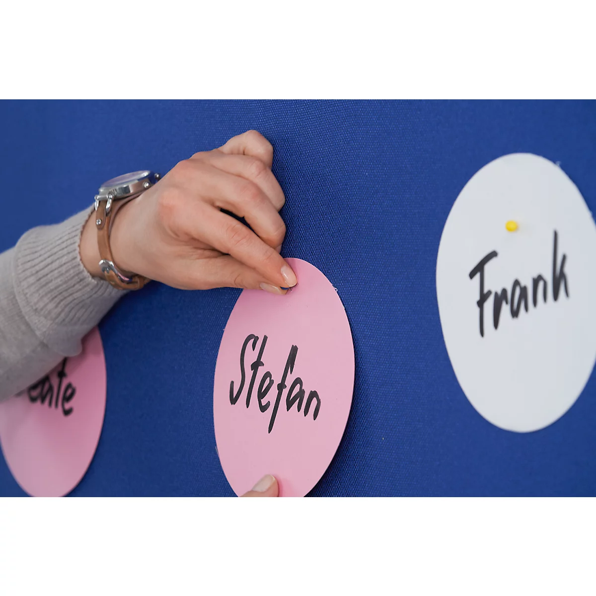 A hand with a wristwatch attaches a pink note with the name "Stefan" to a blue wall. The names "Frank" and "ante" are written on other notes.