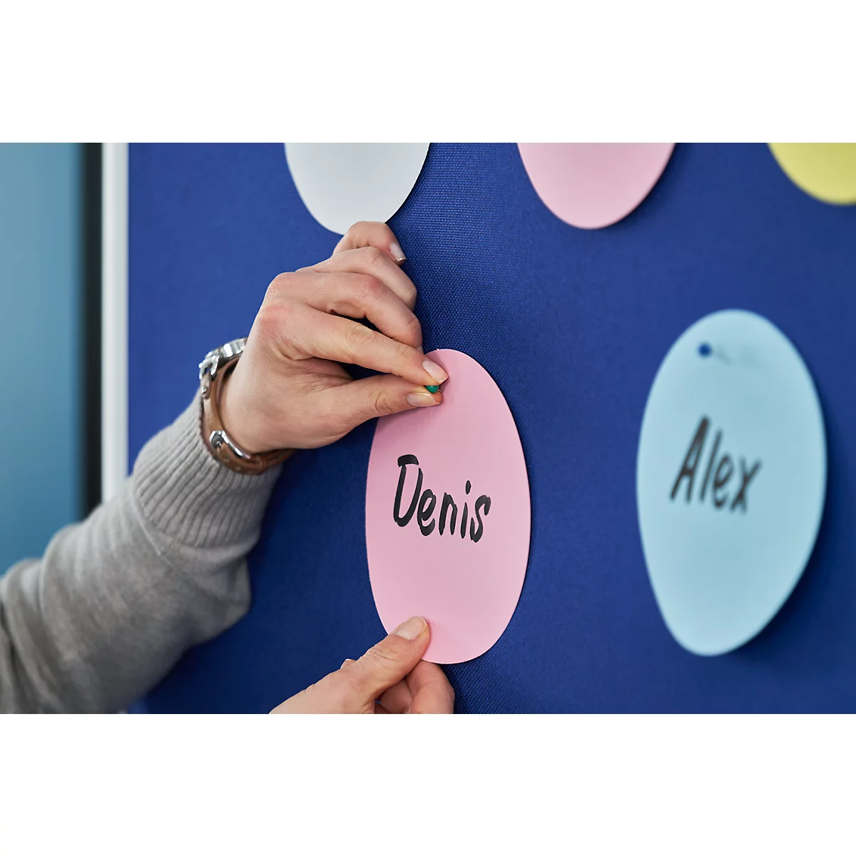 Hand placing a green thumbtack in a pink note with the name "Denis". Blue pinboard in the background.