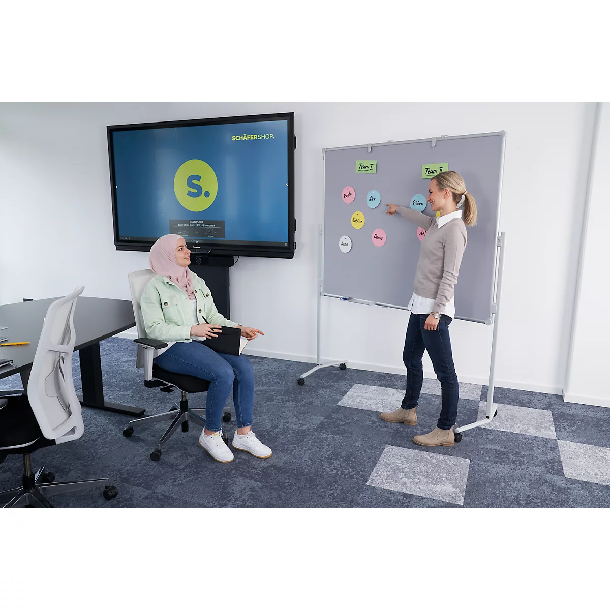 Two women in an office. One woman in a hijab is sitting, the other is standing by a whiteboard with notes.