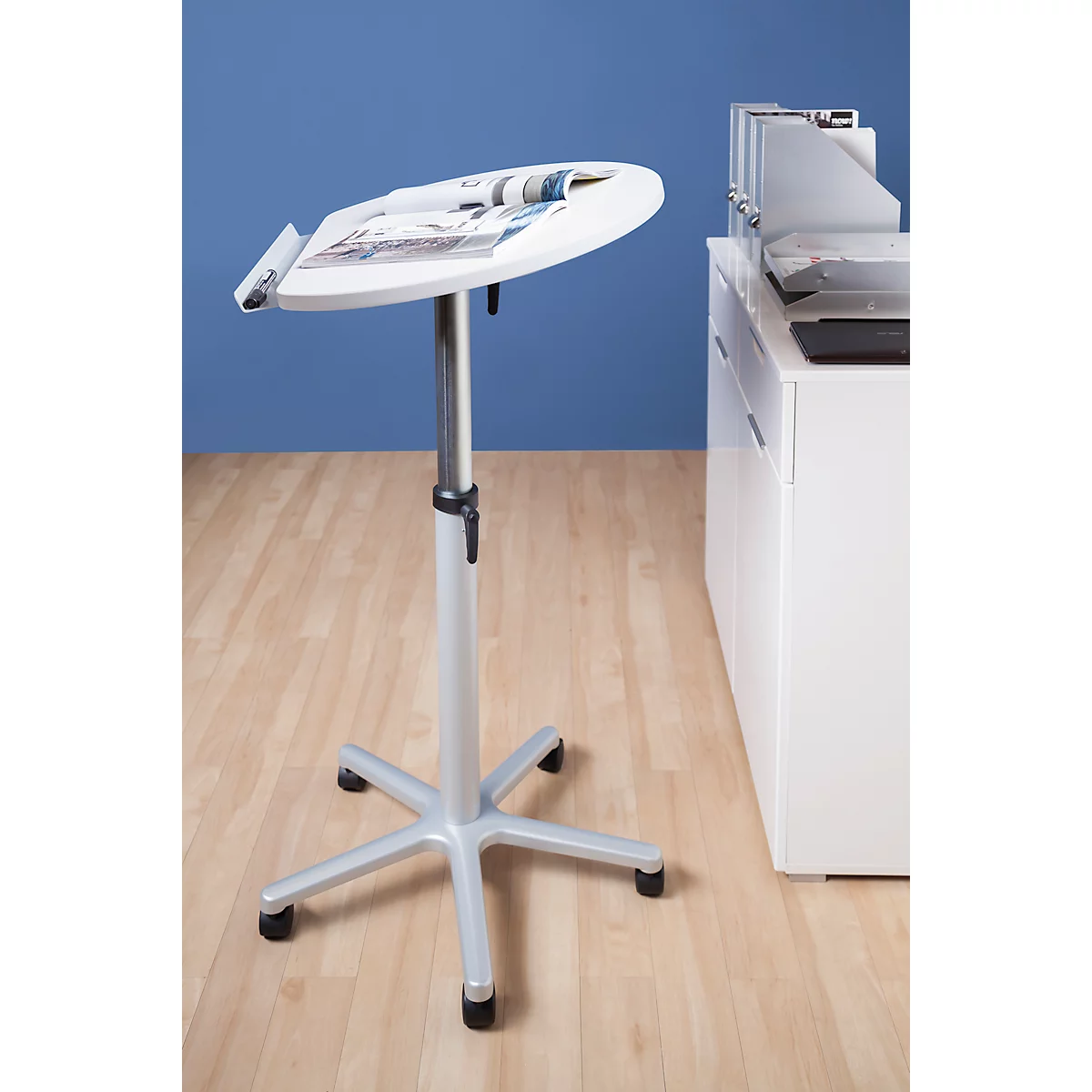 White height-adjustable standing desk with magazine. Stands on light brown wooden floor in front of blue background. White office furniture in the background.
