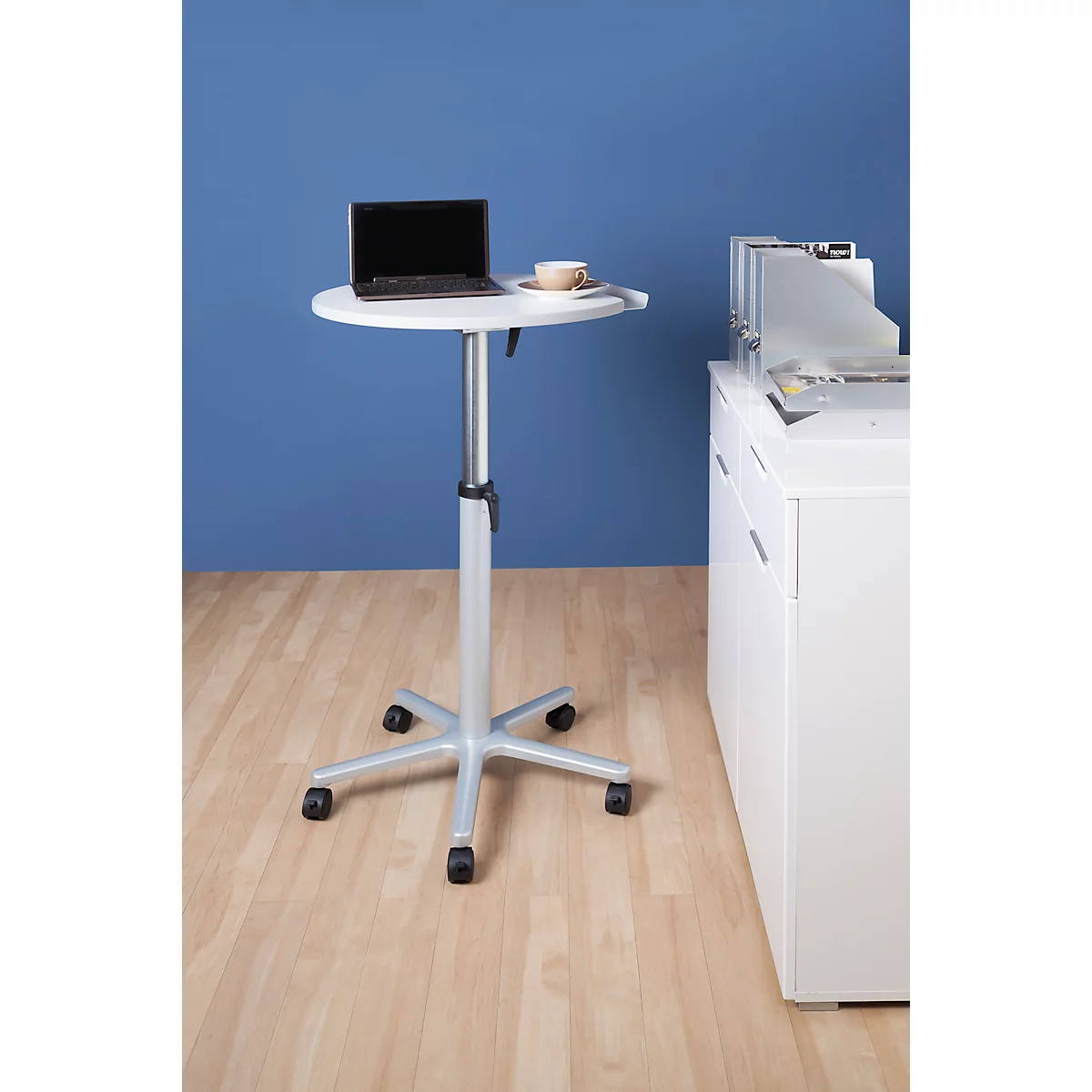 Small, white standing desk with laptop and coffee cup. Wheels on the base. Blue background. Light wood parquet.
